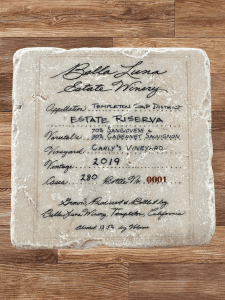 A close-up view of the Bella Luna Estate Winery, 2019 Estate Riserva Marble Coaster. The coaster features an elegant design showcasing the winery's logo and the vintage details. The coaster is made from high-quality marble and is engraved with intricate patterns that reflect the rich blend of Sangiovese and Cabernet Sauvignon grapes from the Templeton Gap District. The design captures the essence of the Estate Riserva, a luxurious varietal with a perfect balance of flavors. The coaster serves as a tasteful reminder of the exquisite wine from Bella Luna's vineyards.